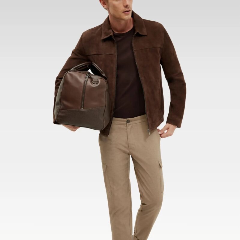 Model wearing the Suede Kael Leather Jacket in dark brown suede goat leather, paired with beige pants and carrying a leather bag